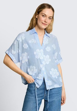 Frau trägt hellblaues kurzärmeliges Hemd mit weißem Blumenmuster, kombiniert mit mittelblauen Jeans, Hand in der Tasche, leicht lächelnd.
