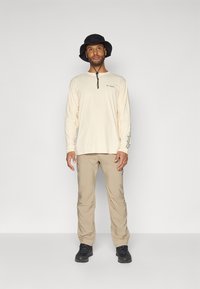 Camicia a maniche lunghe color crema con collo a zip, pantaloni beige e cappello a secchiello nero. L'outfit è completato da calzature nere.