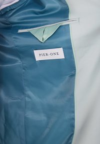 Interiorul unei jachete albastre și verzi deschise, cu un buzunar și o etichetă albă cu inscripția "PIER·ONE". Textură netedă cu un finisaj lucios.