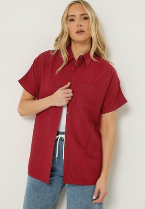 Frau trägt ein locker sitzendes, rotes Kurzarm-Hemd über einem weißen Top und blauen Jeans, steht vor einem einfarbigen Hintergrund.