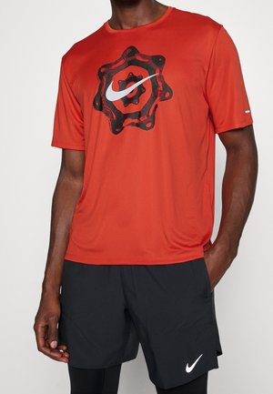 Homme portant un t-shirt rouge Nike avec un motif d'engrenage noir et un logo Nike blanc, associé à un short noir Nike et des leggings noirs.