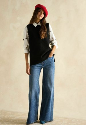 Schwarze Strickweste über einem weißen Hemd mit Tiermotiven, kombiniert mit weiten blauen Jeans und einem roten Barett. Glatte Texturen und lässiges Design.