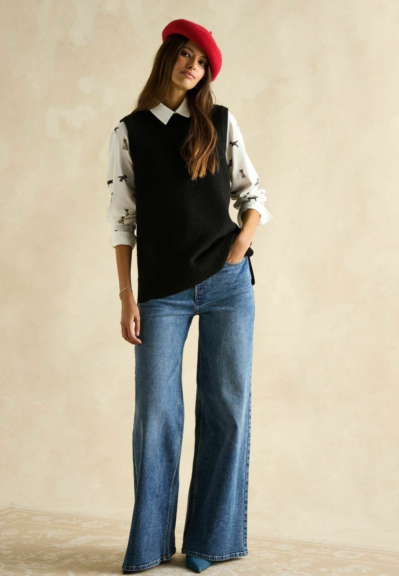 Schwarze Strickweste über einem weißen Hemd mit Tiermotiven, kombiniert mit weiten blauen Jeans und einem roten Barett. Glatte Texturen und lässiges Design.