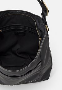 Svart läder tote-väska med en slät yta, som har ett rymligt inre, en dragkedjeficka och en logotagg. Guldiga metallaccenter synliga.