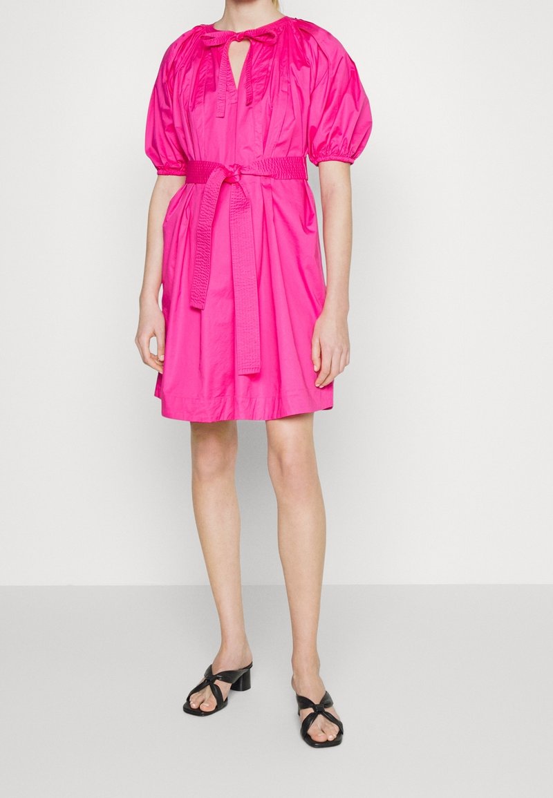 Woman wearing a bright pink knee-length dress with puff sleeves, a waist belt, and black heeled sandals, standing against a plain background.