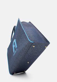 Denimová taška přes rameno s světle modrými detaily a logem. Má trojúhelníkový tvar, pevné rukojeti a zlaté hardware akcenty na dně.