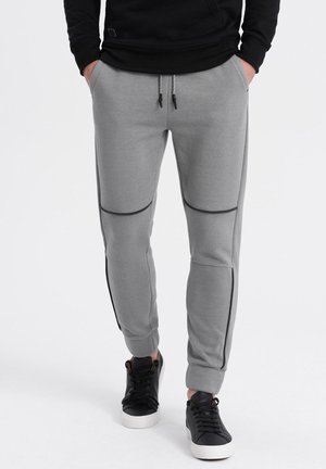 Pantalons de sweat gris avec texture côtelée, dotés d'accents zippés noirs sur la cuisse et les panneaux latéraux, associés à des baskets noires et à semelles blanches.