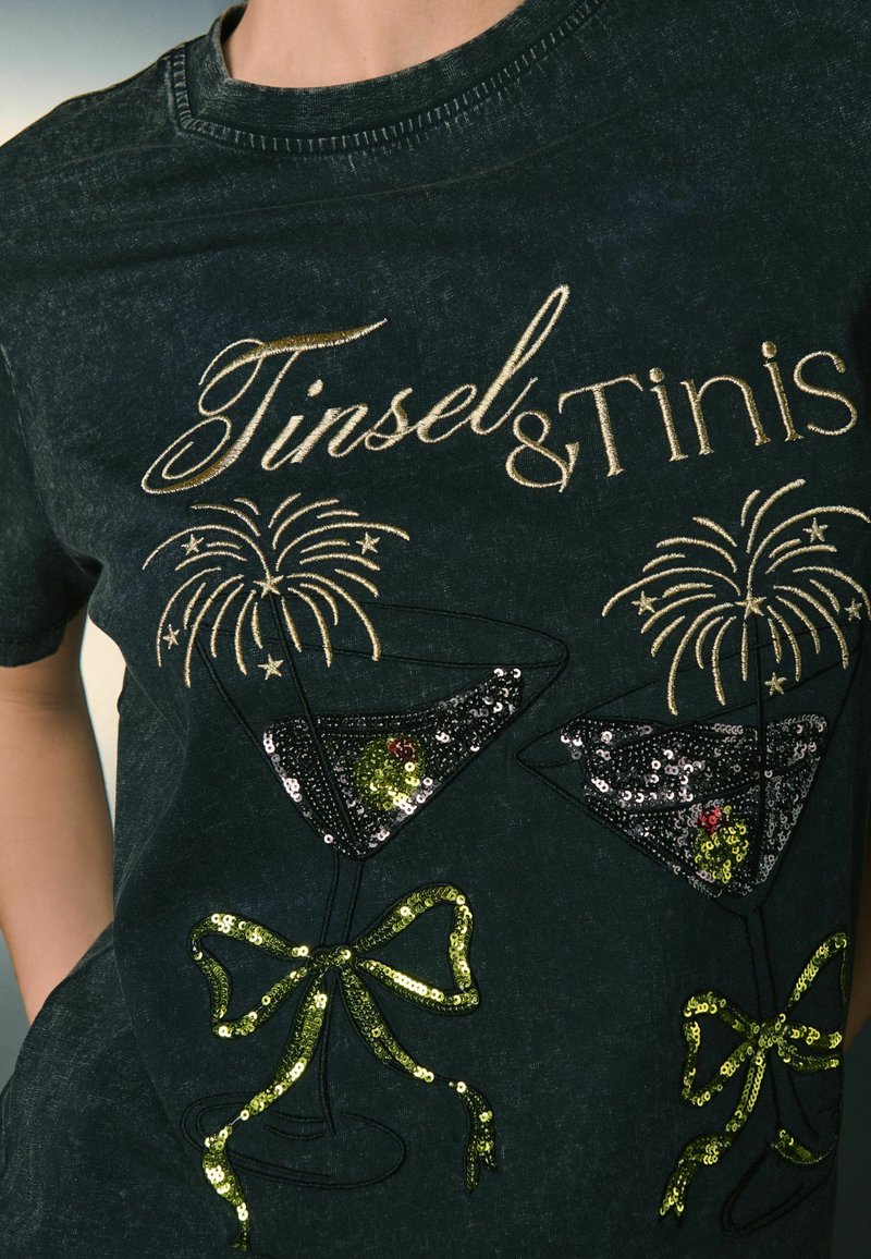 Black T-shirt with metallic gold "Tinsel & Tinis" text. Features sequined cocktail glasses with green sequins, and decorative bows in sequins.