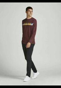 Tricou burgundy cu mânecă lungă, cu un text grafic colorat pe piept, asortat cu pantaloni negri și adidași albi.