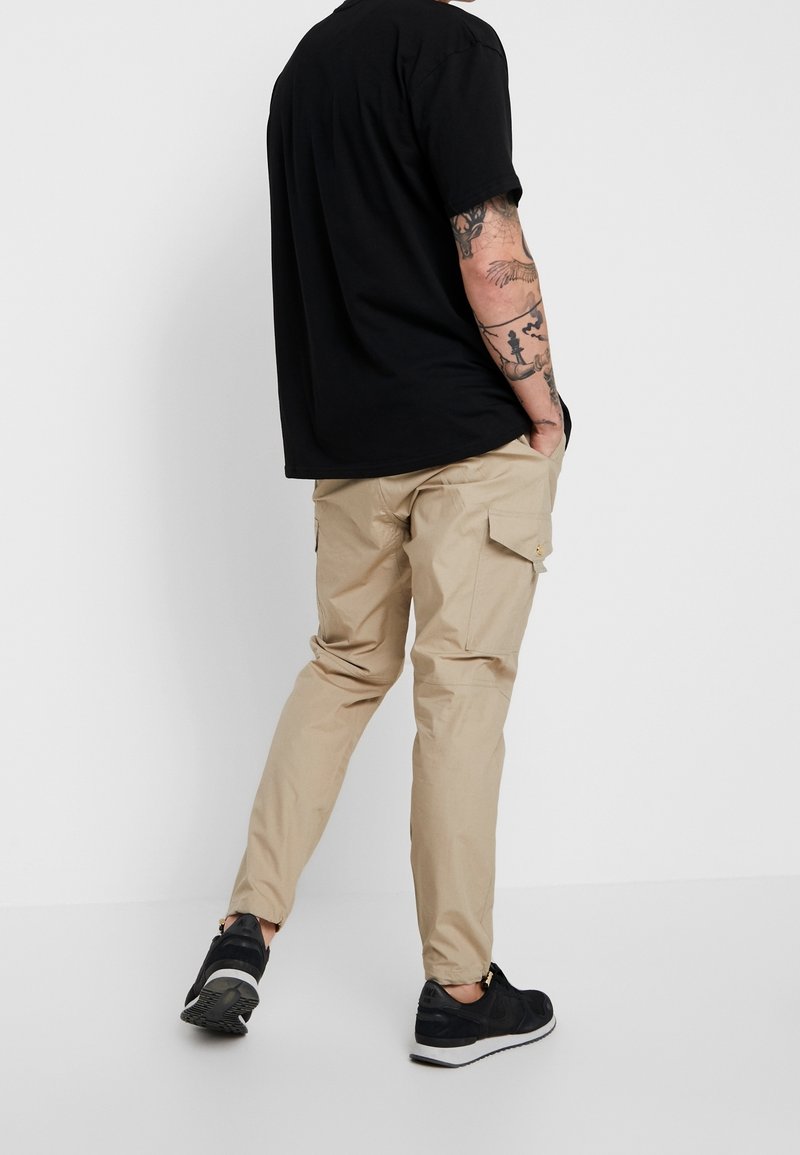 Beige Cargohosen mit zwei Seitentaschen, schmaler Passform und gesteppten Details, kombiniert mit einem schwarzen T-Shirt und schwarzen Sneakers.