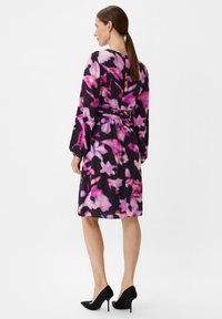 Schwarzes Kleid mit floralem Muster in verschiedenen Rosatönen und Lila. Mit langen Ärmeln, tailliertem Schnitt und fließender Silhouette. Schwarze High Heels.