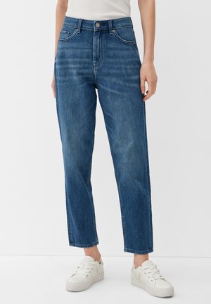 Žena nosící modré džínsy s rovnými nohavicemi a středním pasem a bílé tenisky, stojící před jednobarevným pozadím.