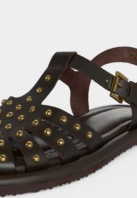 Σανδάλι από σκούρο καφέ δέρμα με χρυσά στρογγυλά τρουκς, λουράκι στον αστράγαλο και χρυσή αγκράφα σε μαλακή σόλα.
