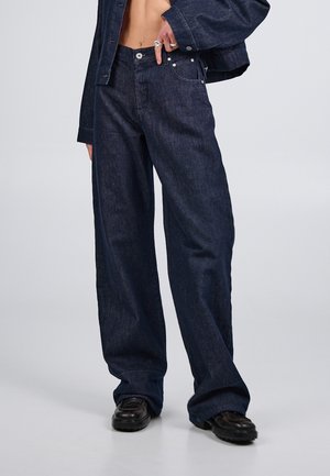 Osoba nosząca ciemne, szerokie dżinsy z szerokimi nogawkami w połączeniu z czarnymi butami oraz dopasowaną, krótką kurtką dżinsową, z ręką wsuniętą do przedniej kieszeni.