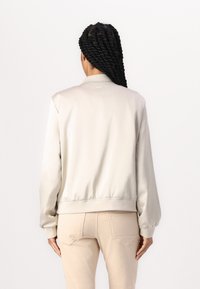 Giacca bomber beige chiaro con una texture liscia e lucida, con polsini e vita a coste. Presenta un design minimalista senza branding visibile.