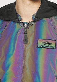 Irydescencyjna kurtka z czarnym kapturem, z zapięciem na zamek błyskawiczny i naszywką "Alpha Industries". Gładka faktura z kolorowym połyskiem.