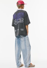 Übergröße schwarzes T-Shirt mit lila Graf design und Text auf dem Rücken, kombiniert mit hellblauen, weiten Jeans und beigen Sneakers.