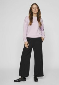 Svijetlopurple dukserica s rebrastim manžetama, uparen s crnim širokim hlačama i lakiranim crnim mokasinama. Glatka tekstura, ležeran dizajn.