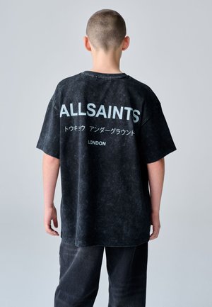Osoba s krátkými vlasy, která nosí černé oversize tričko AllSaints s japonským textem a nápisem „London“ na zádech.