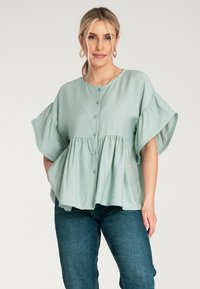 Jasnozielona bluzka z zapięciem na guziki z przodu, szerokimi rękawami i marszczoną talią. Wykonana z miękkiego materiału o luźnym kroju. Połączona z ciemnymi dżinsami.