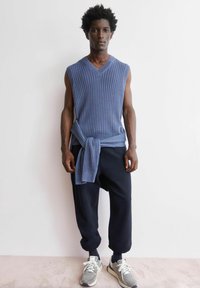 Blauwe geribbelde mouwloze trui met een V-hals, gecombineerd met donkerblauwe broek. Losse silhouet, gestrikt in de taille, afgewerkt met grijze sneakers.