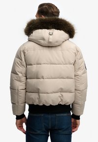 Beżowa kurtka puchowa z futrzanym kołnierzem, pikowany design, elastyczne mankiety i czarny ściągacz u dołu. Posiada detal z logo na ramieniu.