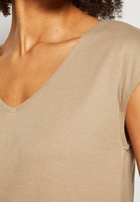 Top beige a V concollo realizzato in tessuto morbido, con maniche corte e una texture liscia. Il colletto è leggermente arcuato.