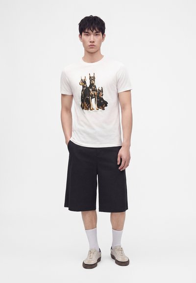 Νέος άνδρας φορώντας λευκό t-shirt με στάμπα τριών σκύλων Ντόμπερμαν, μαύρα φαρδιά σορτς, λευκές κάλτσες και μπεζ αθλητικά παπούτσια, στέκεται μπροστά σε λευκό φόντο.