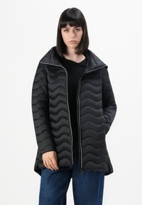 Vrouw met steil donker haar, gekleed in een zwarte golvende gewatteerde ritsjas en blauwe jeans, staand tegen een witte achtergrond.