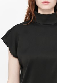 Žena v černém topu bez rukávů s vysokým límcem, volnými rameny a měkkou texturou látky.
