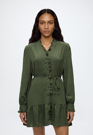 Femme aux longs cheveux foncés portant une robe verte à manches longues boutonnée avec des volants et une ceinture nouée à la taille, debout devant un fond uni.