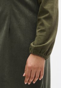 Olívazöld, bordázott ruhaujj gumírozott mandzsettával és finom fényességgel. Kéz semleges körömlakkal, texturált szövet előtt.