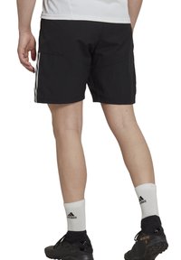 Pantaloni sportivi neri con strisce laterali, in tessuto leggero, con vita elastica e design aderente. Indossati con calze sportive bianche e scarpette nere.