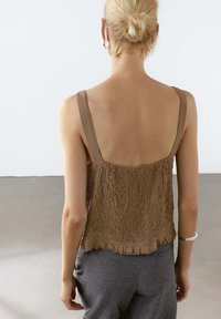 Brown textured crop top with thin straps and fringed hem, paired with gray pants. The fabric has a crinkled appearance and subtle shine.