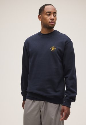 Hombre que lleva una sudadera azul marino con cuello redondo y un pequeño logo circular amarillo en el pecho, y pantalones gris claro, mirando hacia un lado.