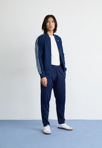 Tengerészkék sportdzseki fehér csíkokkal, tengerészkék sportnadrággal párosítva. Fehér póló alatta és fehér sneaker kék részletekkel.