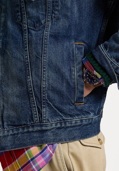 Jeansjakke med klassisk snitt, blå farge, lys falming og gule sømmer. Har en rutete skjorte synlig under og et klokke på håndleddet.