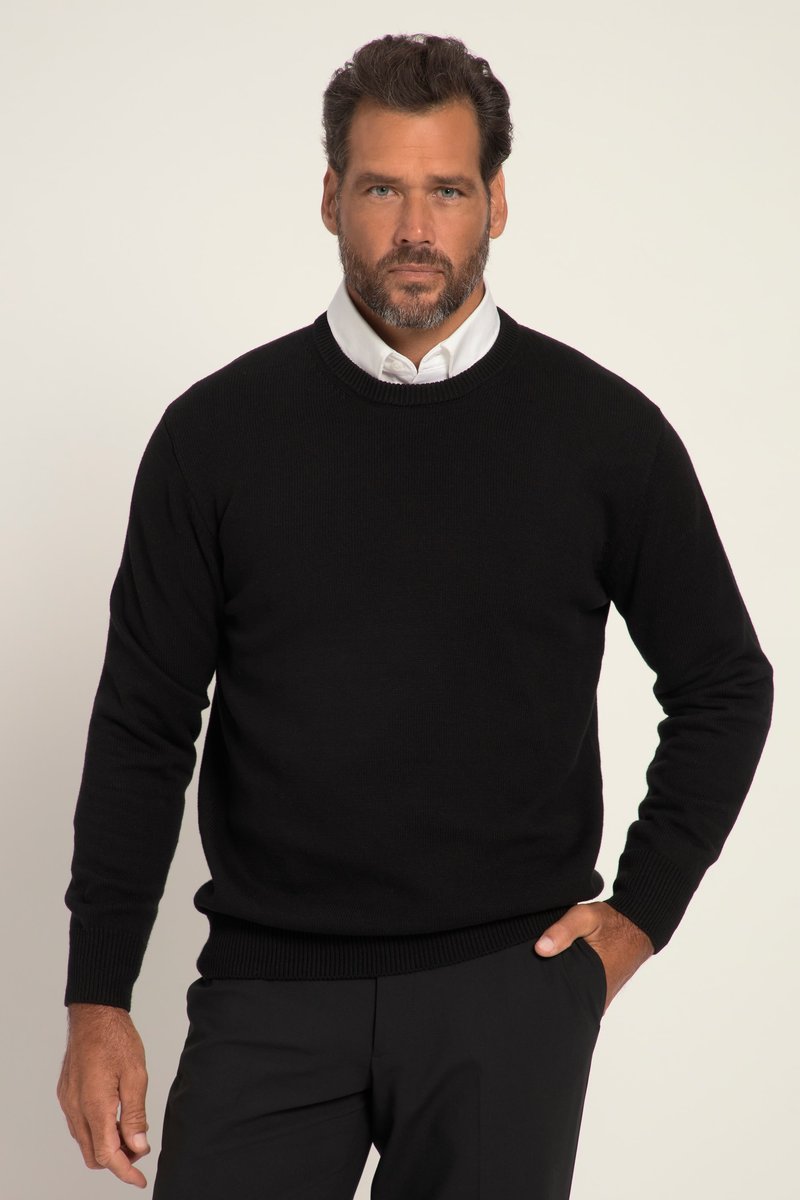 Hombre con barba que lleva un suéter negro sobre una camisa blanca con cuello, de pie contra un fondo claro y liso con una mano en el bolsillo.