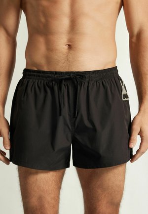 Homme portant un short de bain noir avec une ceinture élastique, un cordon de serrage et un petit décapsuleur attaché à la poche latérale.