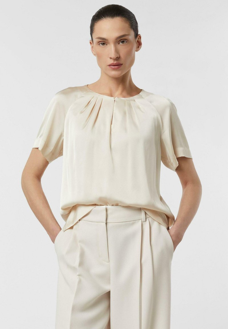 Frau mit zurückgebundenem dunklem Haar, die eine cremefarbene Seidenbluse mit kurzen Ärmeln und passende hochtaillierte Hosen trägt, die Hände in den Taschen.