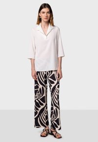 Blouse blanche à col montant avec des manches trois-quarts, associée à un pantalon large au motif de feuilles noir et crème, portée avec des sandales noires.