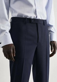 Marineblauwe op maat gemaakte broek met een vlakke voorkant, zijzakken en een knoopsluiting. Gemaakt van een gladde stof met een subtiele glans.