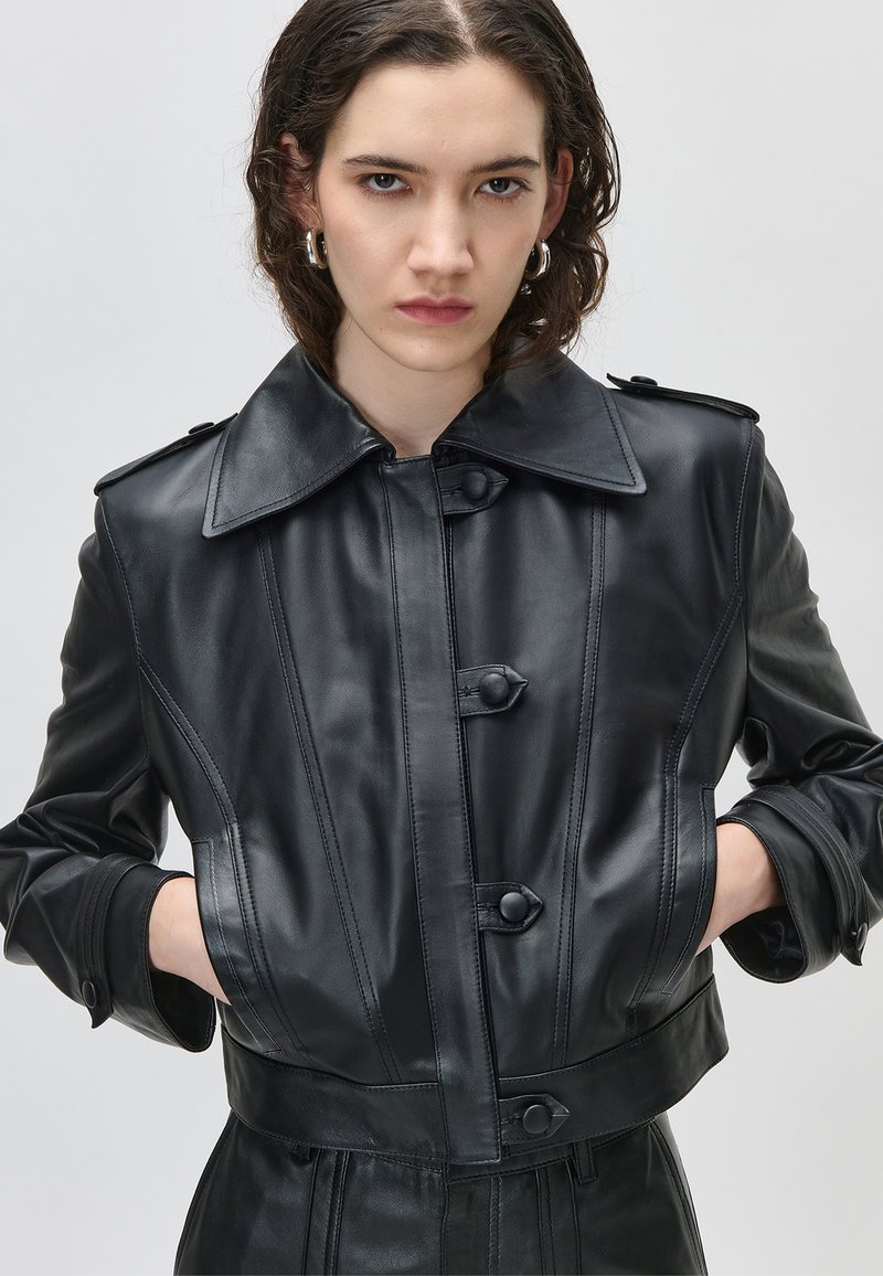 Mlada osoba s kratkom kosom nosi crnu kožnu kratku jaknu s kopčama na dugmad i rukama u džepovima na jednobojnoj pozadini.