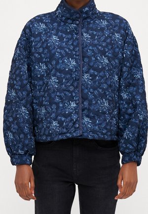 Vatteret jakke i marineblå med blomsterprint, trykknapper foran, høj krave og lange puffede ærmer, båret med sorte bukser.