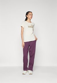 Camiseta crema con un estampado gráfico dorado, combinada con pantalones deportivos morados de corte holgado y zapatillas blancas. Tela suave, estilo casual.
