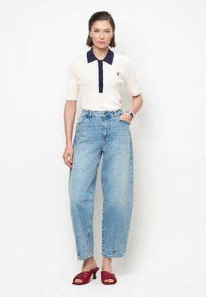 Vrouw draagt een wit poloshirt met korte mouwen en een marineblauwe kraag, lichtblauwe wijde jeans met hoge taille en rode sandalen met hakken, staand met hand in de zak.