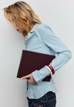 Bordo bilježnica stisnuta uz plavu košulju na pruge, s crvenim i bijelim narukvicama na zapešću. Pozadina je jednobojna i svijetle boje.