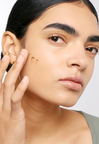 Närbild av ett ansikte med ljusbrun hud, som bär en gyllene ring. Beige produktspår på kinden, glansiga läppar och naturliga ögonbryn.