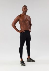 Črne športne pajkice iz raztegljivega materiala z gladko teksturo in zoženim dizajnom. Nošene z temnimi supergami.