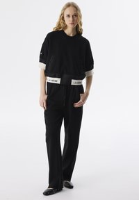 Schwarzer, kurzer Sweatshirt mit einem weißen Logoband, kombiniert mit lockeren schwarzen Hose. Das Outfit besteht aus weichem Stoff und subtilen Akzenten.
